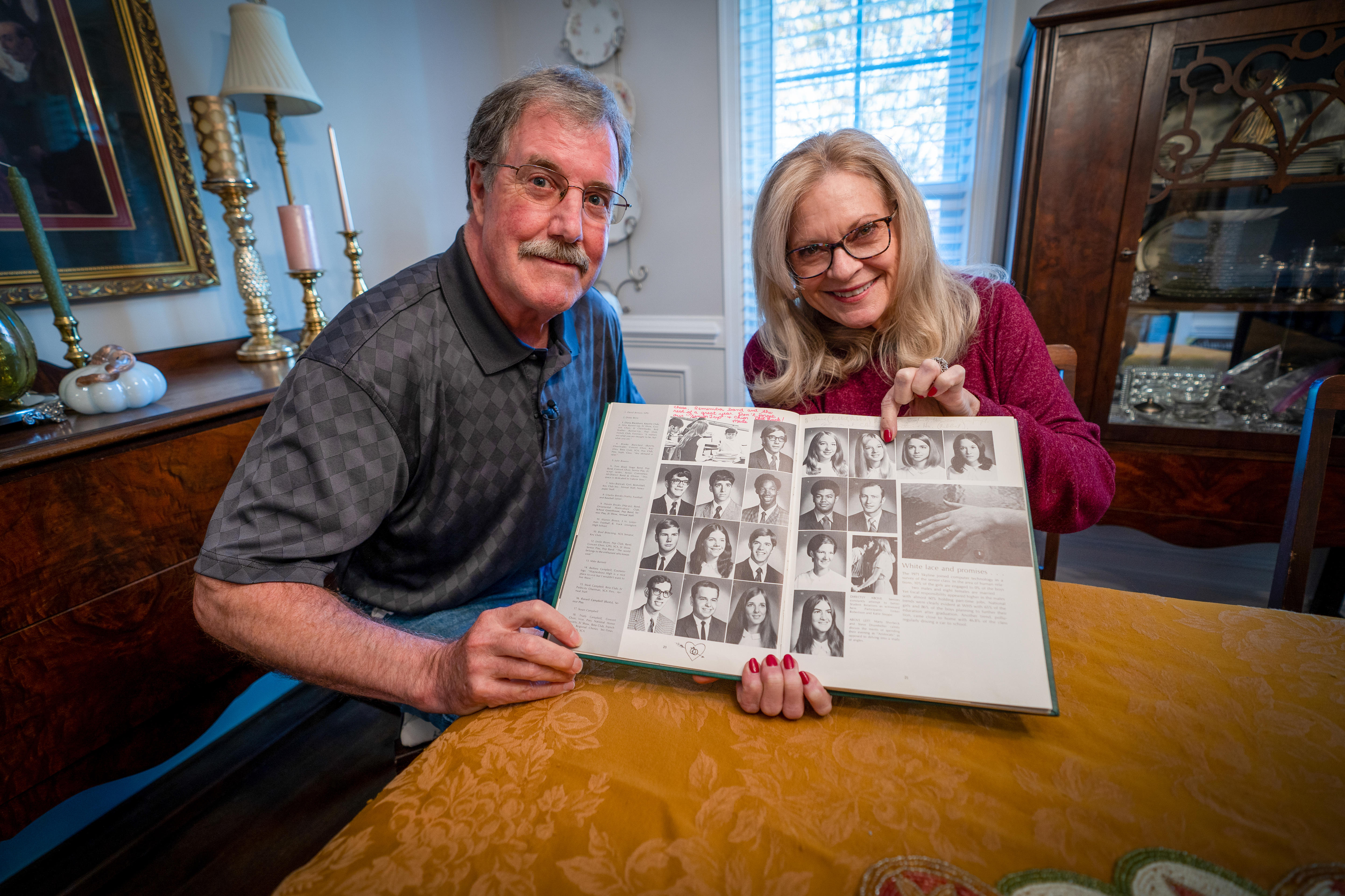 A man and a woman sit at a dining table, holding an old high school year book between them